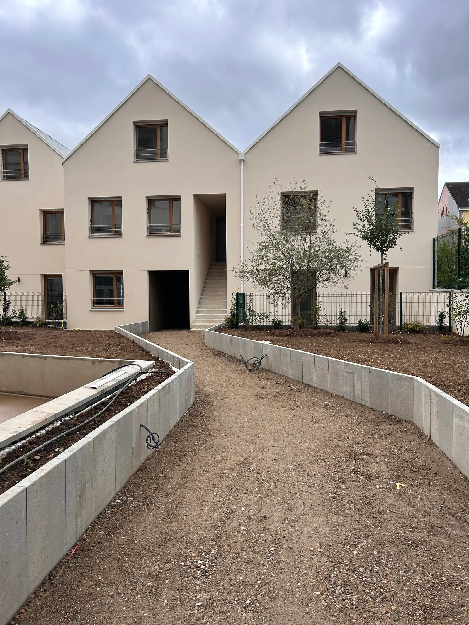 Les maisons à pignons vues depuis le jardin par temps couvert : cheminement en stabilisé, murs de soutènement, jeunes plantations.