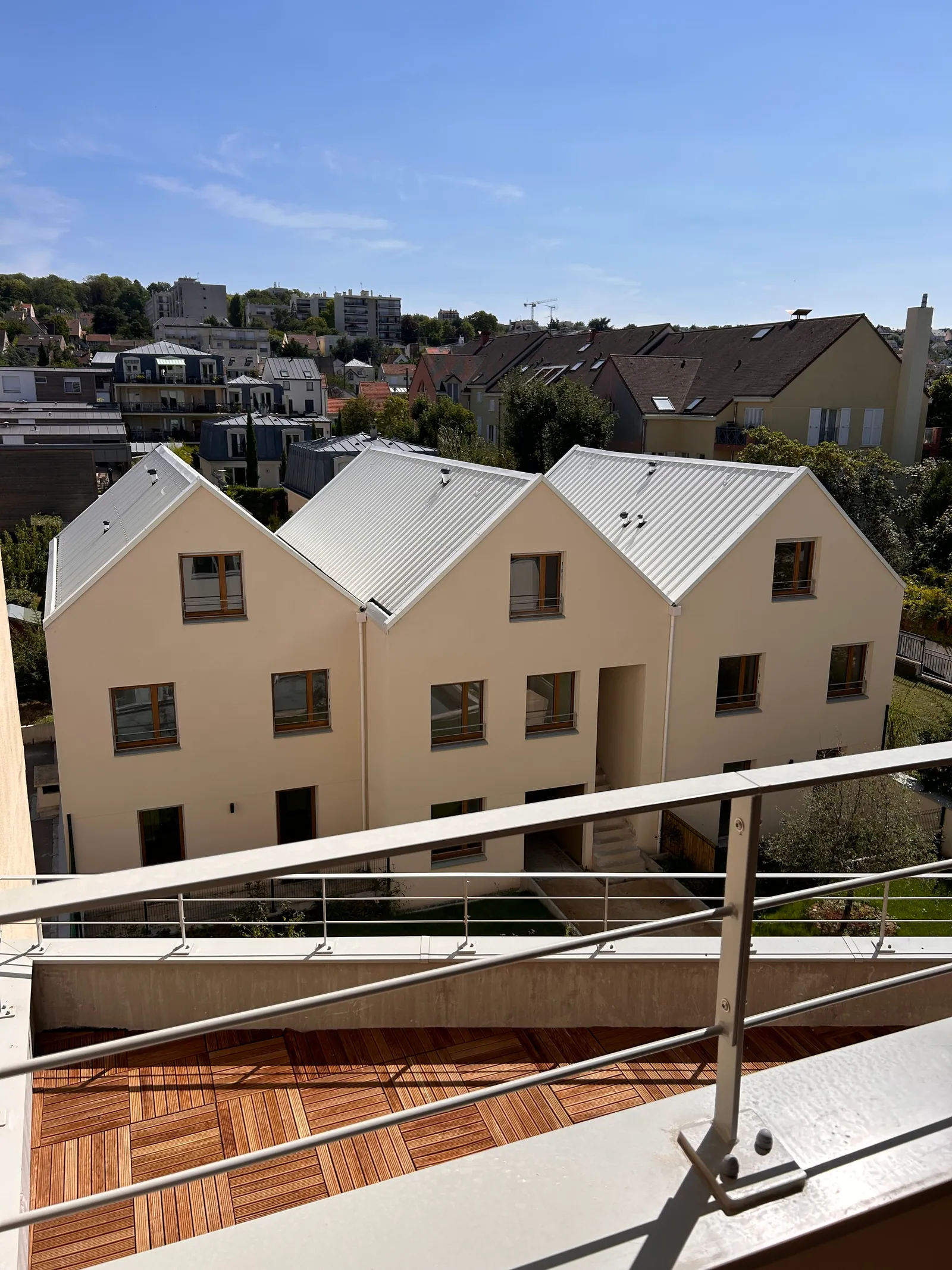 Les toitures en bac acier des maisons à pignons vues depuis un balcon de l'immeuble, avec le platelage bois de la terrasse au premier plan.