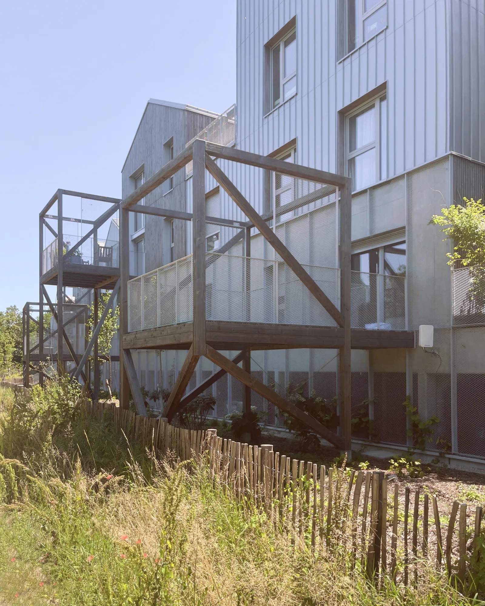 Une terrasse-cabane suspendue sur poteaux bois au-dessus de la prairie, ganivelles et végétation spontanée en pied.