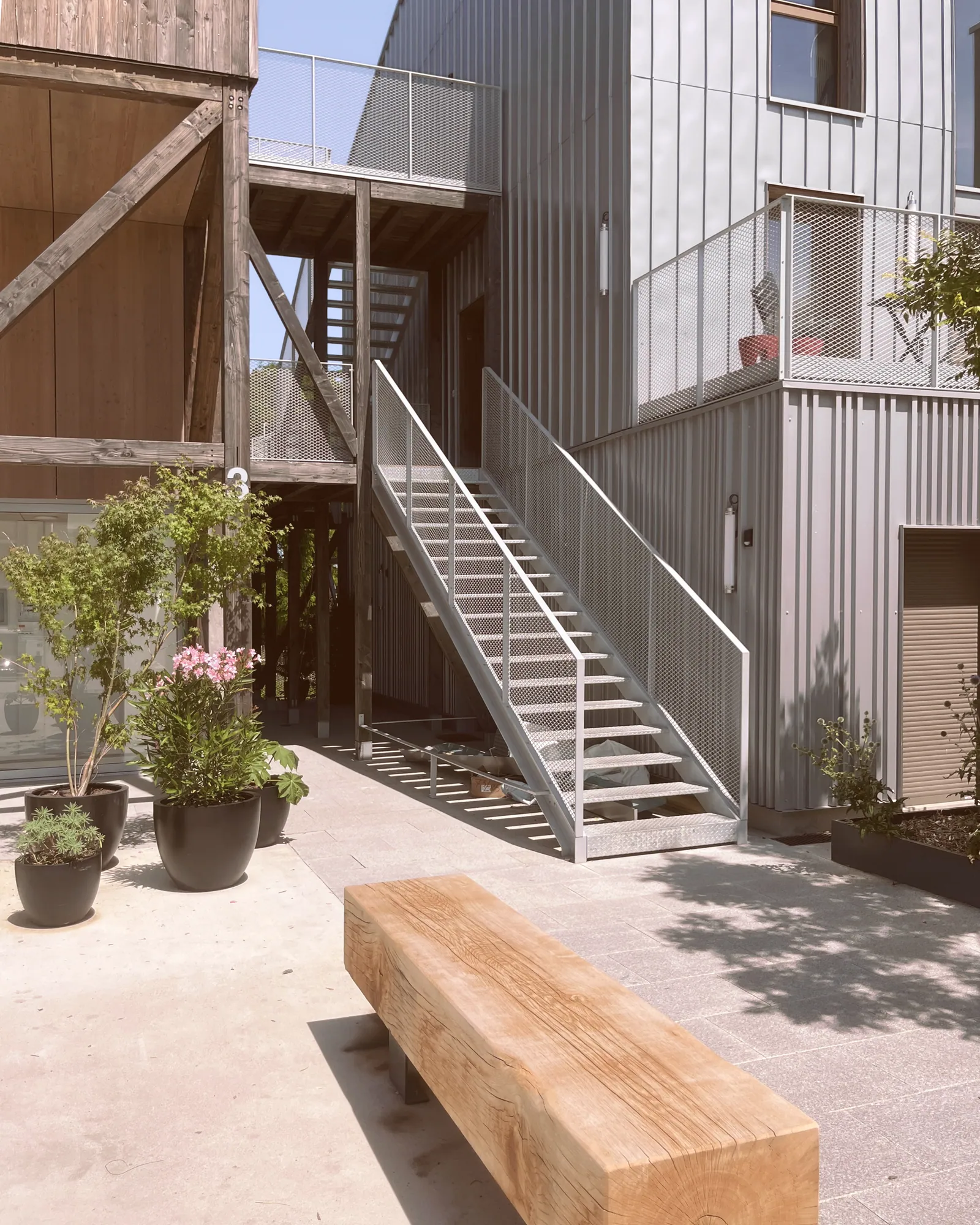 Cour intérieure : banc en bois massif, escalier métallique, plantes en pot au pied des façades bardées.